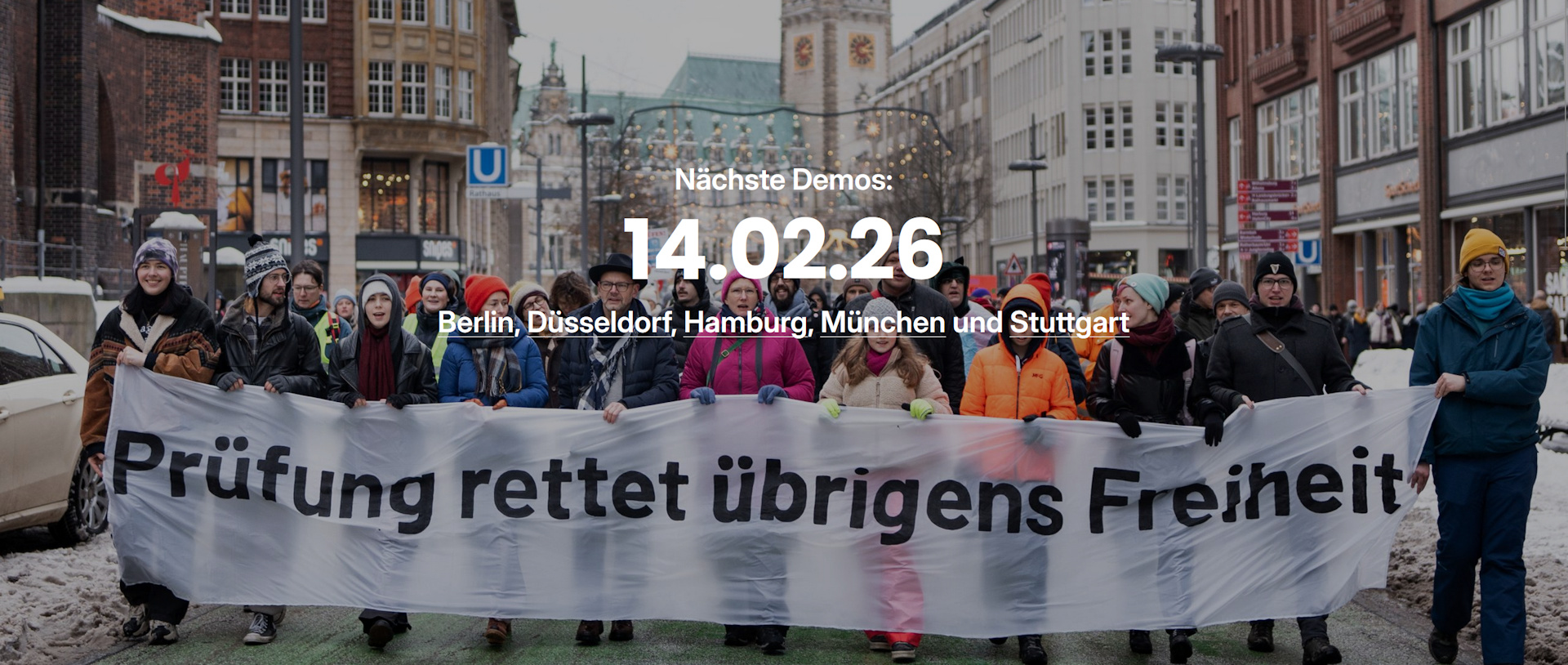 Menschen bei einer Demo mit dem Banner "Prüfen rettet überigens Freiheit", darüber der Hinweis Nächste Demos 14.02.2026 Berlin, Düsseldorf, Hamburg, München und Stuttgart